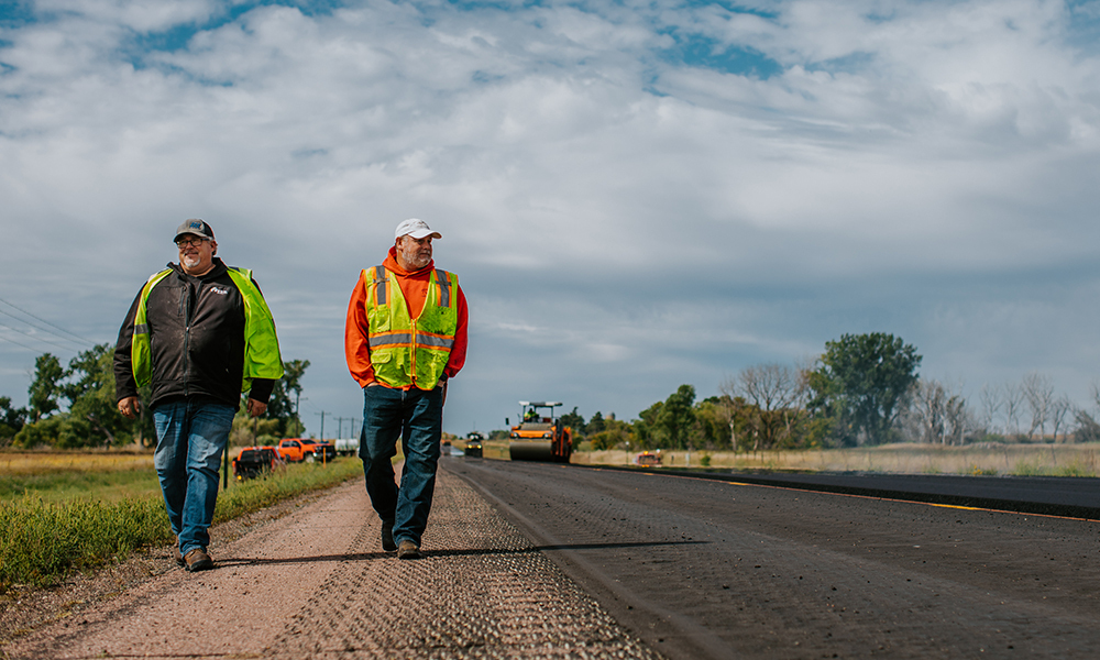 Two road construction crew members walk the side of the highway to inspect their work